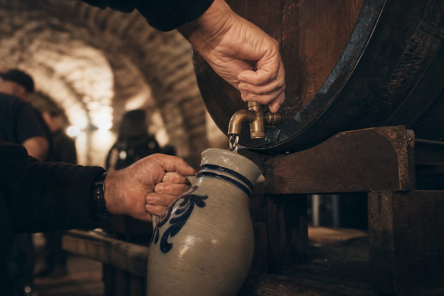 Zwei Hände zapfen Apfelwein aus einem Holzfass in einen grauen Bembel, dahinter ein dunkler Gewölbekeller.