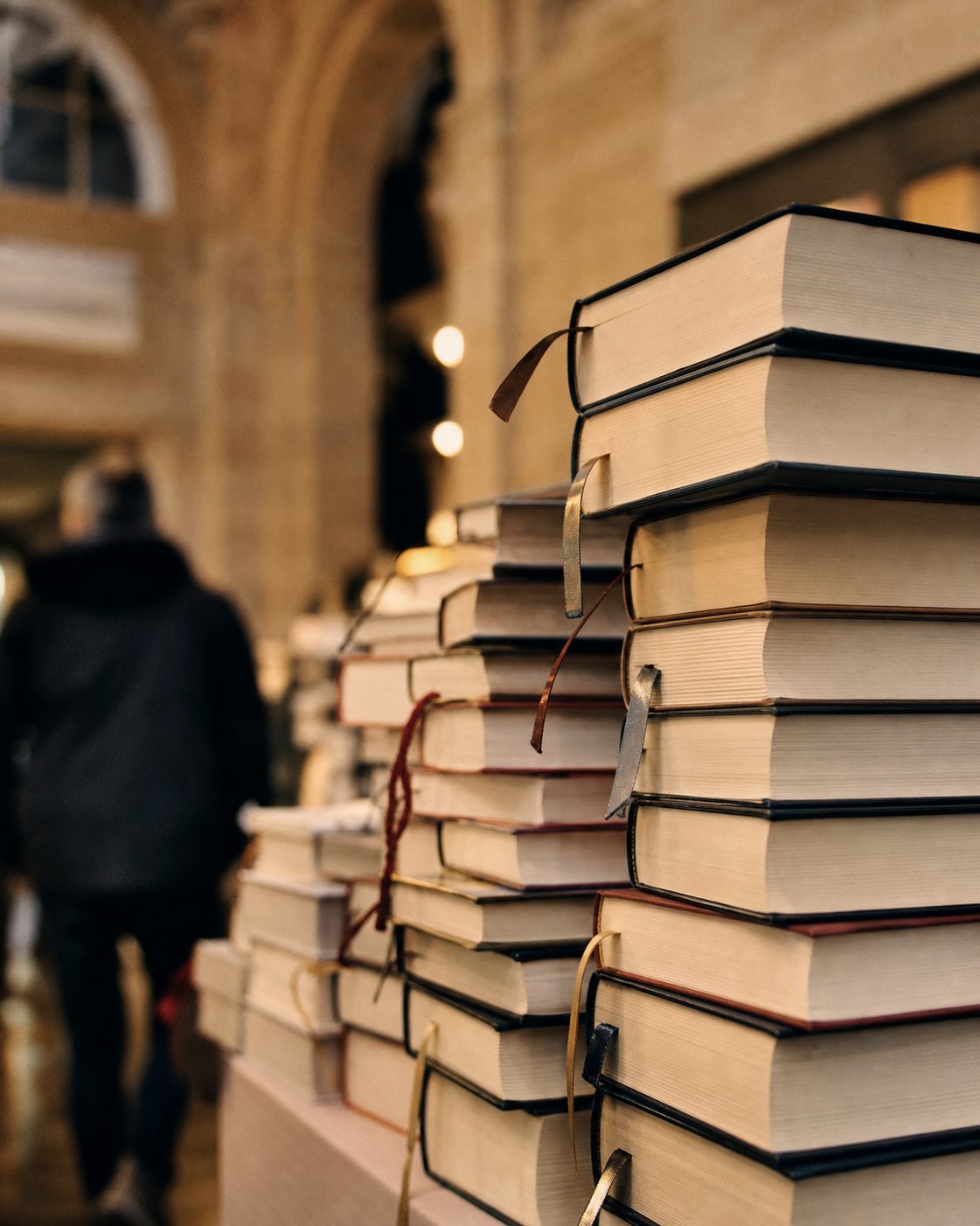 Nahaufnahme gestapelter Bücher mit farbigen Lesezeichenbändern an einem Messestand im warmen Hallenlicht, hochkant.