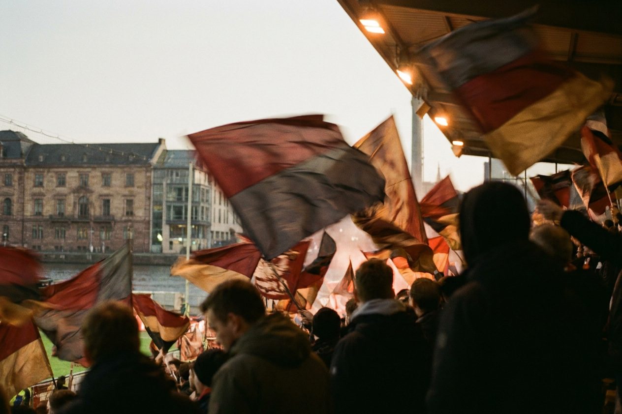 Meer aus geschwenkten Fahnen in einer Fußballkurve bei Flutlicht, verwischte Bewegungsspuren.