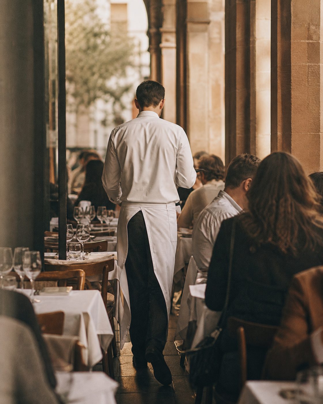 Ein Kellner in weißer Schürze, von hinten gesehen, bewegt sich zwischen den eng stehenden, weiß eingedeckten Tischen des Bistros.