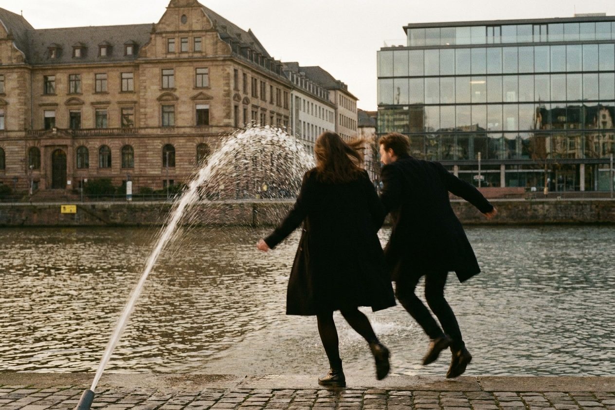 Zwei Passanten springen lachend vor einem Wasserstrahl zur Seite, mit Bewegungsunschärfe und abgewandten Gesichtern.