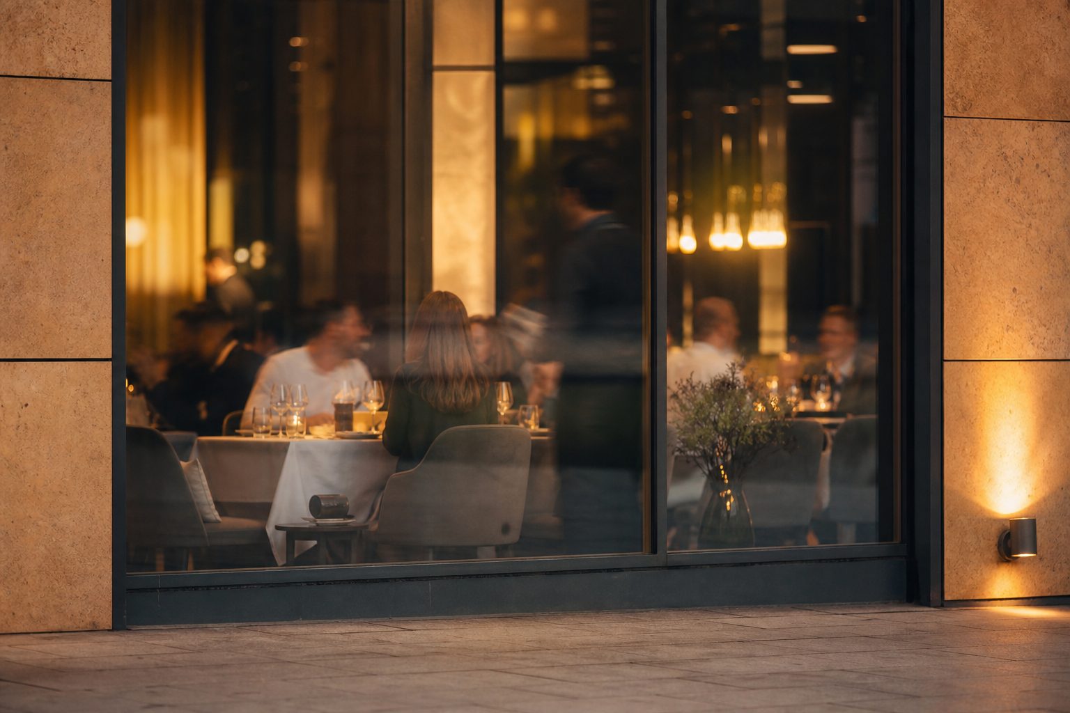 Blick durch eine Schaufensterscheibe auf den ersten Abendservice eines Lokals: Kerzenlicht, gedeckte Tische, Gäste in Unschärfe.