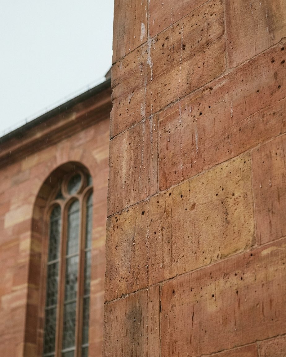 Regennasse rote Sandsteinmauer der Paulskirche mit hohem Rundbogenfenster, warme Steintöne vor blassem Himmel.