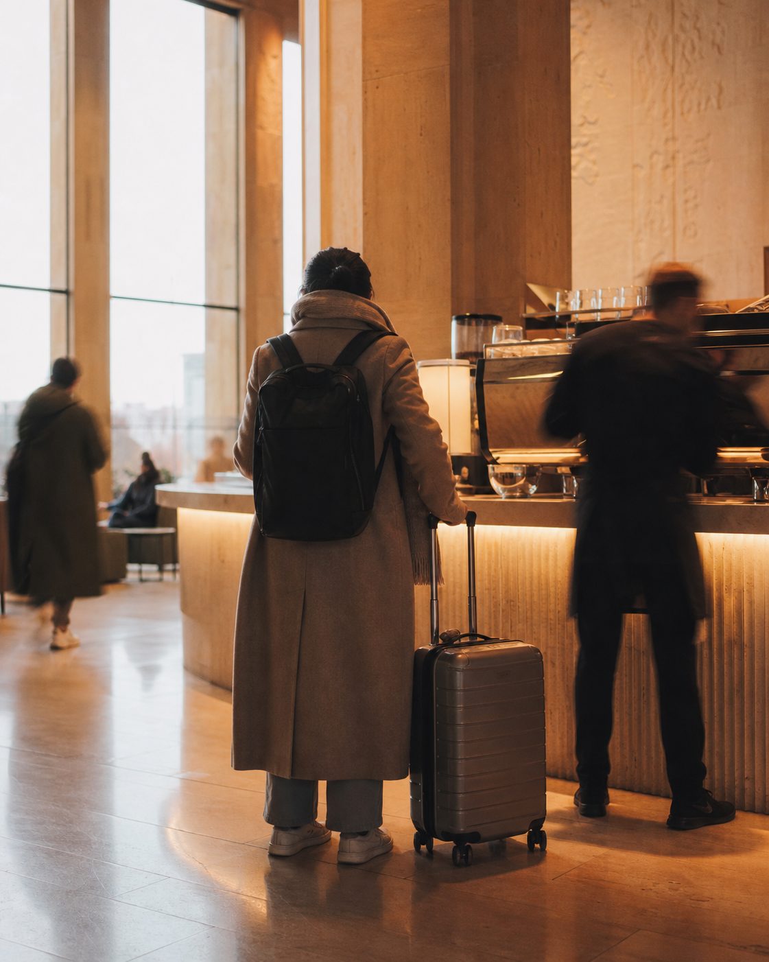 Reisende von hinten mit Trolley an einer hell erleuchteten Kaffeetheke.