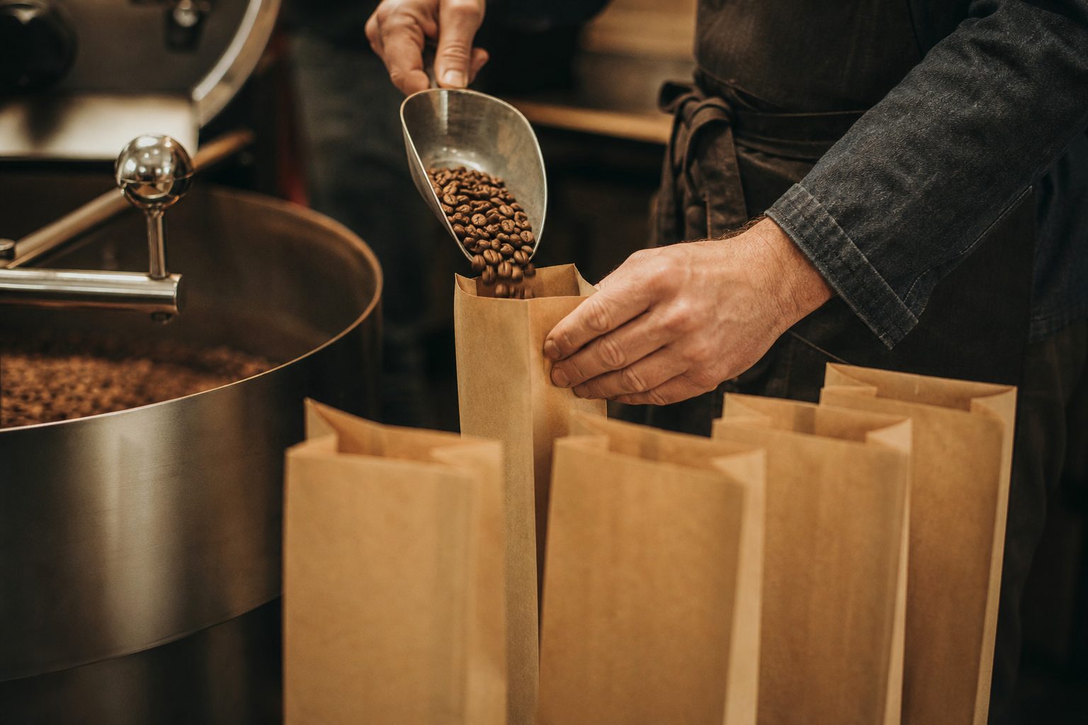 Hände eines Handwerkers füllen frisch geröstete Kaffeebohnen in Papiertüten, das Gesicht bleibt außerhalb des Bildes.