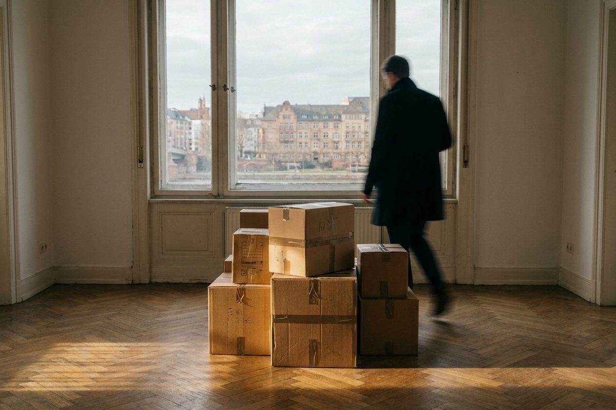 Ein Stapel gepackter Umzugskartons in einem leeren Altbauzimmer, Sonnenstreifen fallen auf das Parkett.